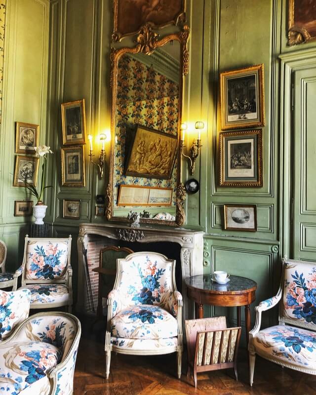 old style furnishings, wooden panelled wall painted light green, decorative fire place with decorative mirror above it and other old style pictures on the walls